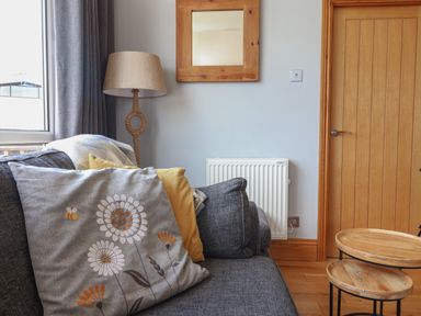 A living room corner with a grey sofa with cushions a floor lamp a wooden framed mirror a radiator a wooden door and nesting wooden tables at Pebble Beach Chalet in Seaton