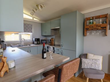 A kitchen with blue cabinets a countertop with a bottle of wine and two wine glasses two brown chairs a gin bar with bottles and glasses on the wall and a gray chair with cushions at Pebble Beach Chalet in Seaton