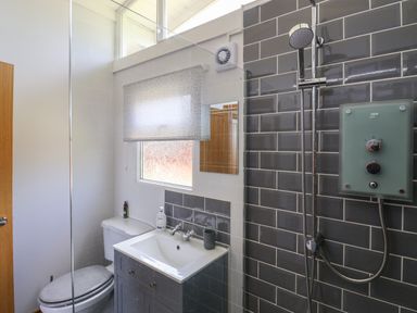 A bathroom with a toilet beside a sink in front of a window and a shower with gray tiles at Pebble Beach Chalet in Seaton
