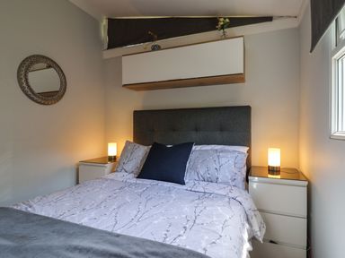 A bedroom with a bed with patterned bedding two bedside tables with lamps a wall mirror and a cabinet at Pebble Beach Chalet in Seaton