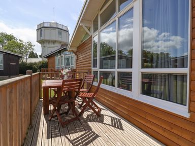 A wooden deck with a table and four chairs outside a cabin with large windows and a tower in the background at Pebble Beach Chalet in Seaton