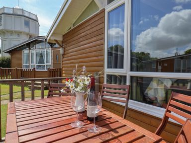 An outdoor wooden table with two wine glasses a bottle of wine and a vase of wildflowers on a patio outside wooden lodges and a white tower in the background at Pebble Beach Chalet in Seaton