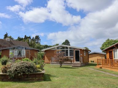 Three wooden chalets with porches surrounded by grass and plants under a partly cloudy sky at Pebble Beach Chalet in Seaton