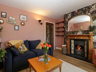 A living room with a navy sofa, wooden coffee table with flowers, fireplace with mirror and floral wallpaper at Holly Cottage in Great Malvern