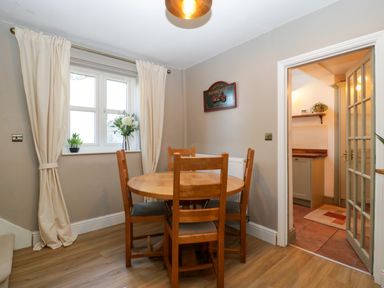 A dining area with a round wooden table and four chairs near a window with curtains and a doorway leading to a kitchen at Holly Cottage in Great Malvern