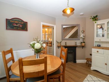 A dining area with a wooden table and chairs a floral arrangement a fireplace with plants and candles a mirror and a white cabinet at Holly Cottage in Great Malvern