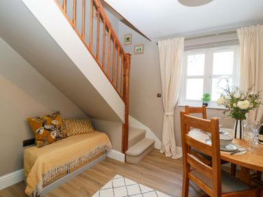 A dining area with a wooden table set for meals beside a window and a small seating area under stairs in Holly Cottage Great Malvern