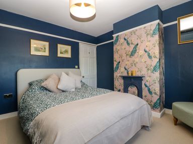 A bedroom with a bed pillows a fireplace with peacock wallpaper and a small green chair at Holly Cottage in Great Malvern