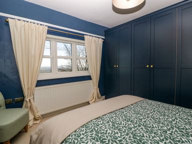 A bedroom with a window and cream curtains a green patterned bedspread a green chair and blue built in wardrobes at Holly Cottage in Great Malvern