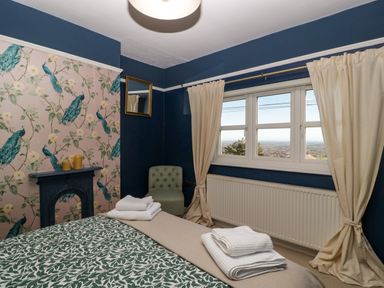 A bedroom with a bed folded towels a chair window with cream curtains and a decorative fireplace at Holly Cottage in Great Malvern