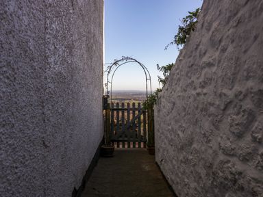 A narrow passageway with white textured walls leading to a wooden gate with an arch and a landscape view beyond