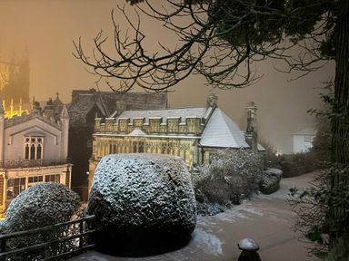 A snow-covered garden path with trimmed bushes and old stone buildings in the background at Holly Cottage in Great Malvern