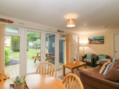 A living room with a brown sofa two armchairs wooden coffee and dining tables and glass doors opening to a garden at 245 Riverside Lodge in Caeathro