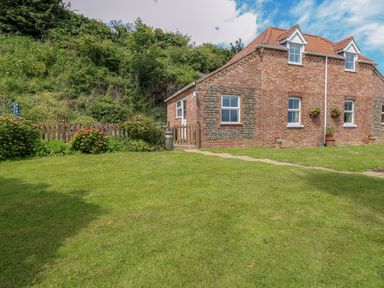 A brick house with tiled roof and white framed windows beside a garden with grass flowers and bushes at Dairy Cottage in West Caister near Caister-on-Sea