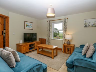 A living room with two blue sofas a wooden coffee table a television on a wooden stand and a window with floral curtains at Dairy Cottage in West Caister near Caister-on-Sea