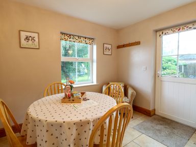 A dining area with a round table covered by a tablecloth and wooden chairs next to a window and door at Dairy Cottage in West Caister near Caister-on-Sea