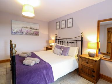 A bedroom with a bed with purple blanket and pillows wooden bedside tables with lamps and framed pictures on the wall at Dairy Cottage West Caister near Caister-on-Sea