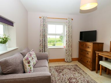 A small living room with a gray sofa floral cushions a wooden dresser with a television floral curtains and a wooden table at Dairy Cottage in West Caister near Caister-on-Sea