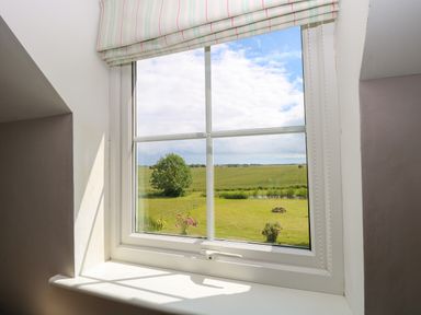 A window with a striped blind overlooking a green field with a tree and picnic table at Dairy Cottage in West Caister near Caister-on-Sea