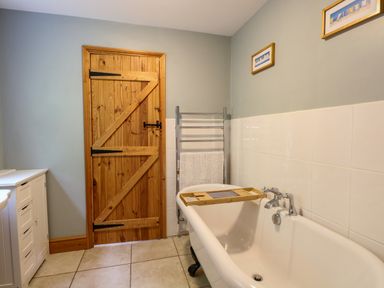 A bathroom with a wooden door a white bathtub with a wooden bath tray a towel rack and framed pictures on the wall at Dairy Cottage in West Caister near Caister-on-Sea