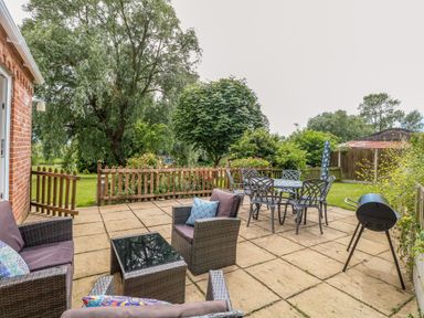 A patio with outdoor seating furniture a metal dining table with chairs a barbecue grill and a wooden fence with trees in the background at Dairy Cottage in West Caister near Caister-on-Sea