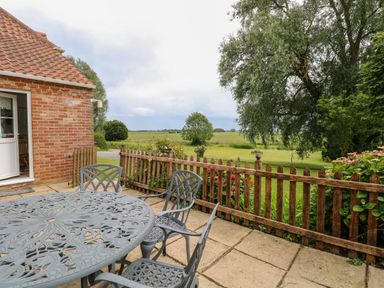 A patio with a metal table and chairs next to a wooden fence and a grassy field at Dairy Cottage in West Caister near Caister-on-Sea