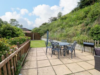 An outdoor patio with a metal table and chairs a wooden fence and greenery at Dairy Cottage in West Caister near Caister-on-Sea