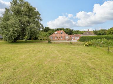 A large grassy lawn with a tree and a brick house with a red roof in the background at Dairy Cottage in West Caister near Caister-on-Sea
