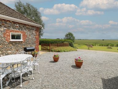 An outdoor patio area with white metal table and chairs gravel ground two flower pots stone wall building and open field at Flint Cottage in West Caister near Caister-on-Sea