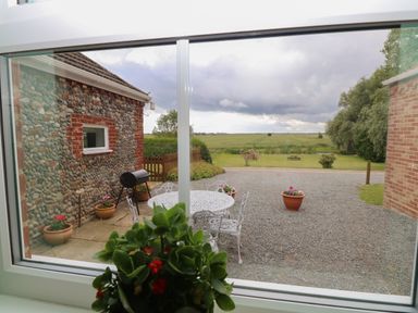 A view of a gravel patio with a white metal table and chairs a barbecue and potted plants outside a flint wall building and a grassy field beyond at Flint Cottage in West Caister near Caister-on-Sea