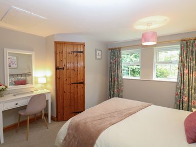 A bedroom with a bed a dressing table with a chair and lamp wooden door and two windows with floral curtains at Flint Cottage West Caister near Caister-on-Sea