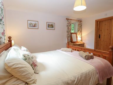 A bedroom with a wooden bed, white bedding, floral pillows, two framed pictures, a wooden dresser with a mirror, and floral curtains at Flint Cottage in West Caister near Caister-on-Sea