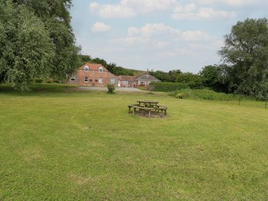 A lawn with a circular wooden picnic table and benches and brick buildings in the background at Flint Cottage in West Caister near Caister-on-Sea