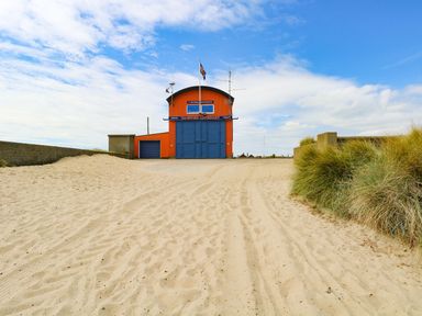 A sandy pathway leading to an orange lifeboat station with blue doors and grassy dunes on the side at Flint Cottage in West Caister near Caister-on-Sea