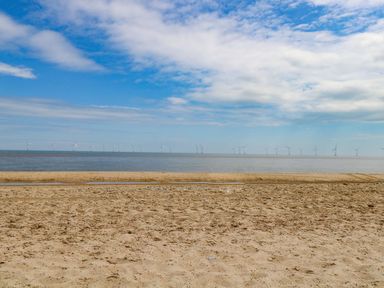 A sandy beach with the sea and a row of wind turbines on the horizon under a partly cloudy sky at Flint Cottage in West Caister near Caister-on-Sea