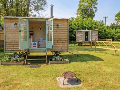 Two wooden shepherd huts on wheels with open double doors on a grassy area with flower beds and a fire pit at Acres Meadow in Maiden Newton