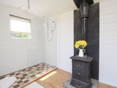 A shower area with a tiled floor and glass partition next to a black stove with a white jug of yellow flowers at Acres Meadow in Maiden Newton