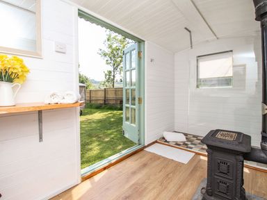 An interior room with a small stove wooden floor a shelf with towels and flowers and a door open to a garden at Acres Meadow in Maiden Newton