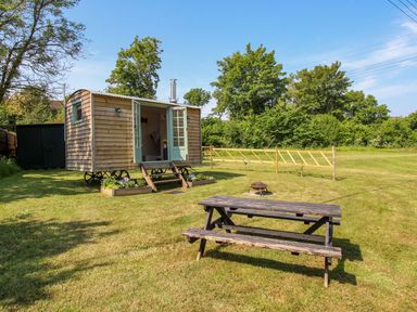 A wooden hut with open double doors on a grassy lawn with a picnic table and fire pit at Acres Meadow in Maiden Newton