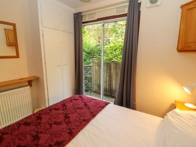 A bedroom with a bed covered with a red patterned blanket, white built-in wardrobes, a window with gray curtains, a wooden shelf, and a small bedside lamp at Bluebells in Chapel Stile