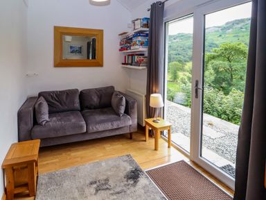 A small living room with a gray sofa two wooden side tables a lamp a wall mirror shelves with books and board games and glass doors overlooking greenery at Bluebells in Chapel Stile
