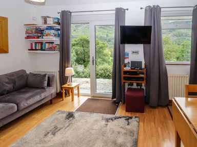A living room with a gray sofa a small lamp on a wooden side table a wall-mounted tv shelves with board games and books and a door and window with gray curtains showing green trees outside at Bluebells in Chapel Stile