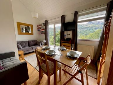 A dining table set with bowls and glasses next to a living area with sofas and a wall-mounted television at Bluebells in Chapel Stile