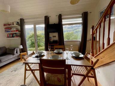 A dining area with a wooden table set for four next to a window and glass door with curtains in a room with a staircase and a sofa