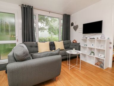A living room with a gray sofa and yellow cushions a white storage unit with a television and a small white round table at Number 24 The Park Kilkhampton