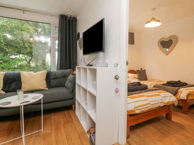 A living room with a gray sofa and a white table next to a bedroom with two single beds at Number 24 The Park in Kilkhampton