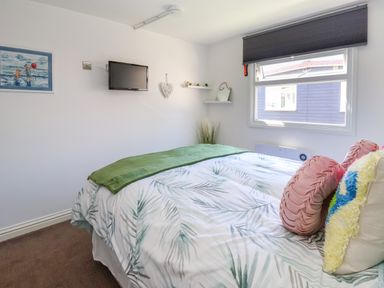 A bedroom with a bed with pillows and a green throw blanket a wall-mounted TV a window with a blind and wall shelves at Sunny South Shore in Bridlington