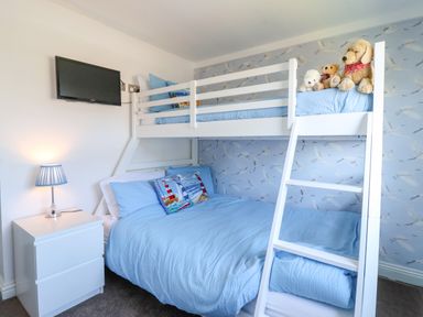 A bedroom with a white bunk bed with blue bedding a bedside table with a lamp and a wall-mounted TV at Sunny South Shore in Bridlington