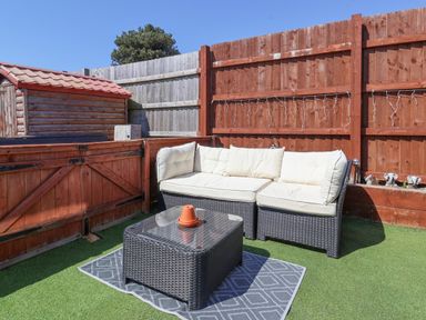 An outdoor seating area with a black wicker couch with white cushions a matching table with a flower pot on top a small shed and wooden fences at Sunny South Shore in Bridlington
