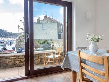 A dining area with a wooden chair and table with a polka dot tablecloth and flowers in a vase next to a large glass door overlooking boats and a waterfront at 7 Island Quay in Salcombe